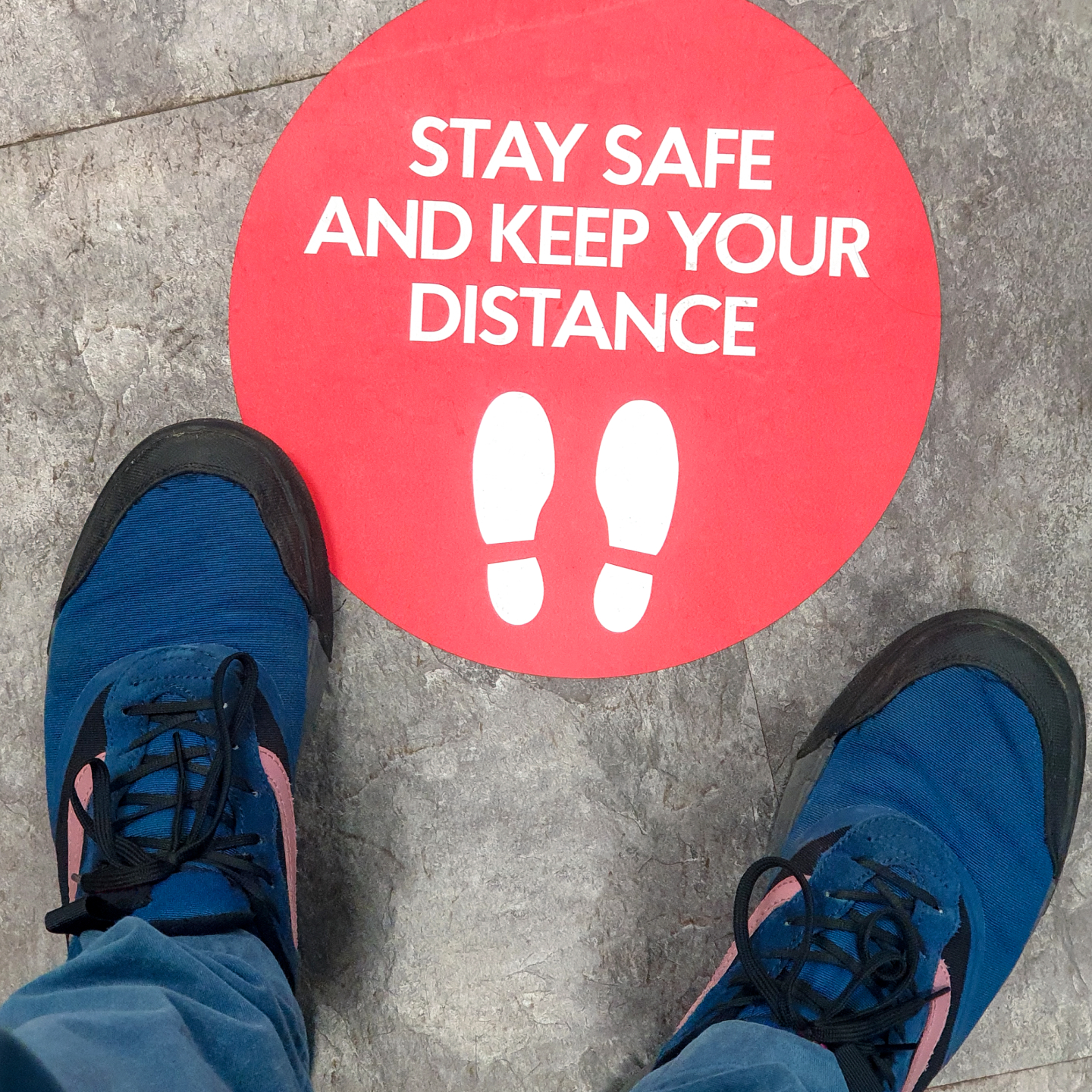 Feet next to a floor sign that says “stay safe and keep your distance&quot;