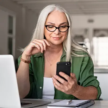 An older woman in her home in front of a laptop and looking at her cell phone